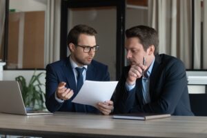 A lawyer and their client in suits sitting at a table next to each other, with the lawyer holding a piece of paper and discussing Massachusetts labor laws for salaried employees.