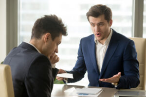An angry male employer sits at his desk and gestures with his hands, presenting a paper to his employee, who holds his head and wonders what to do if your employer is retaliating against you.
